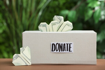 Box with word DONATE and dollar banknotes in shape of heart on table outdoors