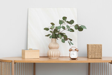 Vase with beautiful eucalyptus branches, clock and decor on table near white wall, closeup