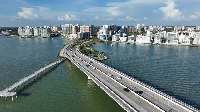 Aerial view of Sarasota, Florida, showing a scenic coastal bridge, waterfront buildings and a boat speeding through clear blue water on a sunny day. Wide shot.