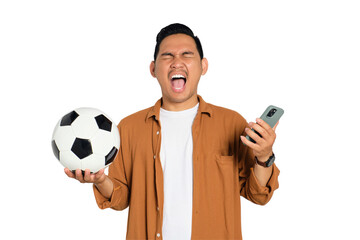Excited young Asian man in casual shirt holding soccer ball and smartphone, shouting yes,...