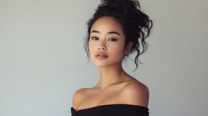 Portrait of a young Asian woman with dark hair in a messy bun, wearing a black off-the-shoulder top against a light grey background.