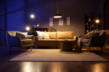 Interior of stylish living room with cozy sofa, coffee table, armchairs and glowing lamps at night