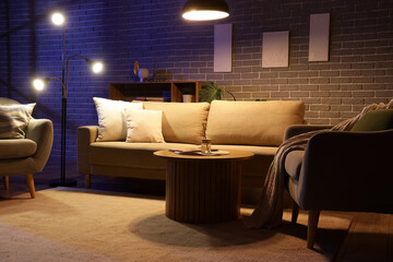 Interior of stylish living room with cozy sofa, coffee table, armchairs and glowing lamps at night