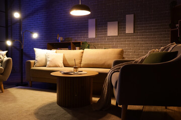 Interior of stylish living room with cozy sofa, coffee table, armchair and glowing lamps at night