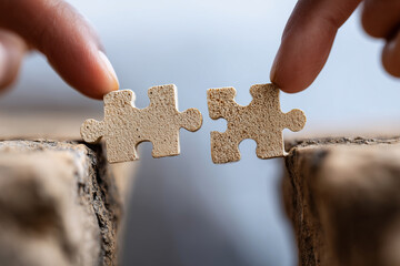 Close up, Two people on opposite sides of a chasm trying to connect bridge puzzle pieces, visual metaphor for bridging the gap, collaboration, overcoming barriers