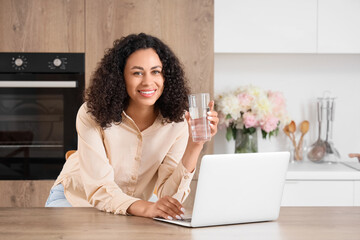 Young African-American woman with glass of water using laptop at table in kitchen