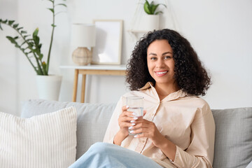 Young African-American woman with glass of water sitting on sofa at home