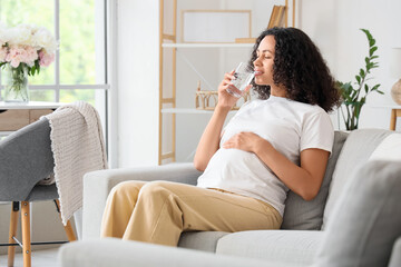 Young pregnant African-American woman drinking water on sofa at home