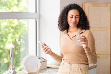 Young African-American woman with glass of water using mobile phone in kitchen