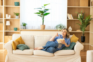 A joyful woman munches on popcorn on her couch, happily interacting with her phone amidst a...
