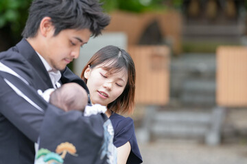 In June 2025, a young Japanese couple in their early twenties is at a Shinto shrine in Aichi. The mother gently helps the father dress their one-month-old baby in a traditional ceremonial kimono.