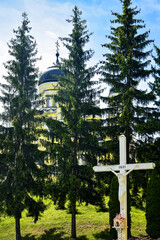 Obraz premium Christian Orthodox white cross with sculpture of Jesus Christ and lamp against green fir trees, big temple and blue sky. Religious theme, faith concept.