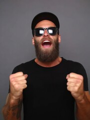 Man in black tee cap  shades clenches fists mouth open against a gray backdrop Tattooed arms are visible
