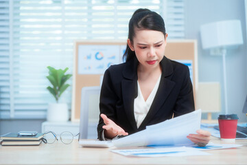 An Asian woman in a formal black suit sits at her office desk, holding documents with a tense...