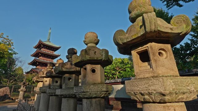 A traditional Japanese pagoda stands prominently behind a row of ancient stone lanterns in Ueno Park, Tokyo. The clear sky and serene setting highlight the timeless beauty of this historical landmark.