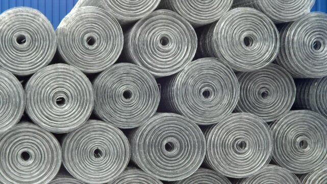 A close-up, tightly framed view of numerous rolls of galvanized wire mesh stacked in an industrial warehouse or storage facility.