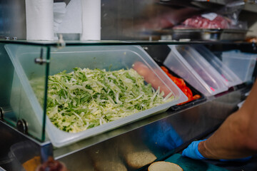 A food service worker wearing blue gloves prepares a sandwich next to bins of shredded lettuce, tomatoes, and other fresh ingredients in a commercial kitchen.