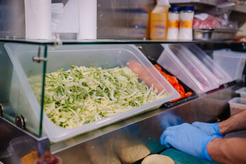A food prep station with shredded lettuce, tomatoes, sauces, and a worker in blue gloves preparing...