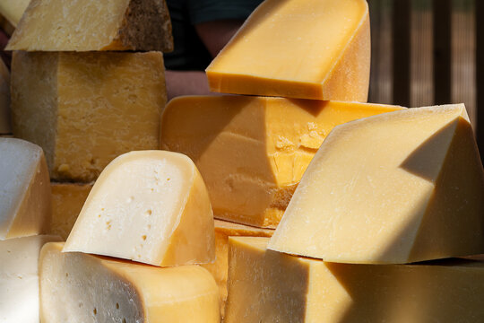 Various types of cheese blocks arranged on a stall, showcasing a variety of textures and colors. The cheeses are stacked and ready for selection by market-goers.