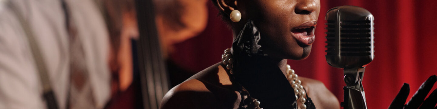 African American female vocalist singing into vintage microphone wearing jewelry, performing jazz music on stage with Caucasian man playing upright bass in background, red curtain behind musicians