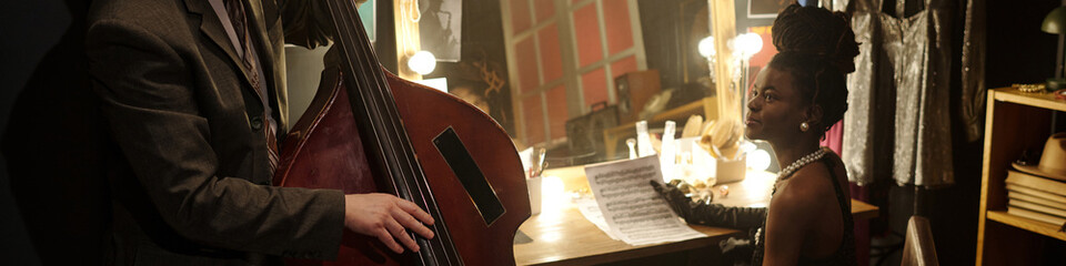 Black young woman at dressing table holding sheet music looking at Caucasian middle aged man playing double bass in backstage area before jazz performance