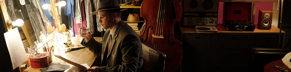 Middle aged Caucasian man wearing hat sitting at dressing room table holding glass and reading sheet music with double bass and audio equipment, preparing for jazz performance
