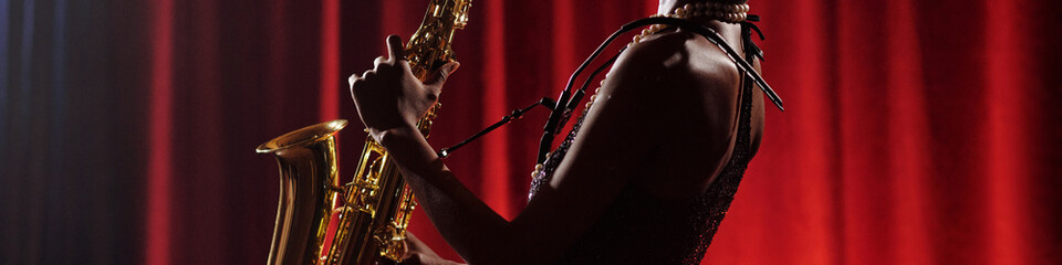 Black young woman playing saxophone on stage in front of red curtain wearing necklace holding...
