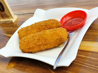 Crispy golden rissoles with savory and delicious filling, served on an elegant white plate, accompanied by a small bowl filled with bright red chili sauce and a fork, on the menu at an outdoor cafe.
