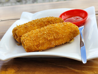 Crispy golden rissoles with savory and delicious filling, served on an elegant white plate, accompanied by a small bowl filled with bright red chili sauce and a fork, on the menu at an outdoor cafe.