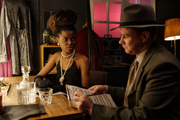 Young Black woman wearing jewelry and middle aged Caucasian man in suit holding sheet music sitting...