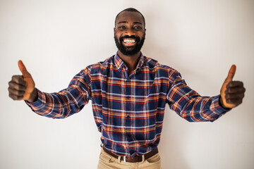 Happy black man in cozy shirt shirt standing in front of wall and smiling. He is showing thumbs up.