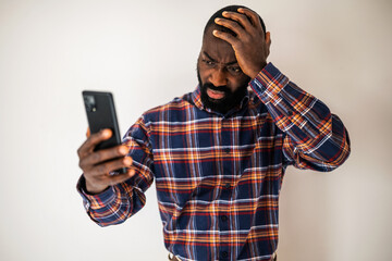 Anxious black man in cozy shirt standing in front of wall and having video call on smartphone.