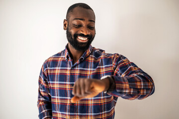 Cheerful black man in cozy shirt standing in front of wall and looking at his wristwatch.