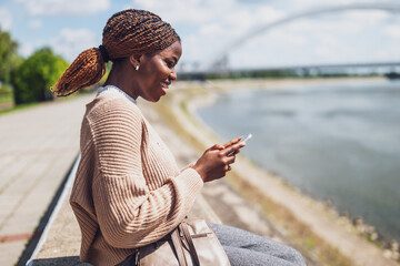 Young happy black woman is using smartphone. She is sitting by the river in the city.
