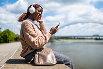 Young black woman is using smartphone and listening music on headphones, outdoor by the river.