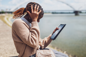 Young anxious black woman is using digital tablet. She is sitting by the river in the city.