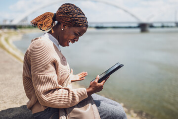 Young stressed black woman is using digital tablet. She is sitting by the river in the city.