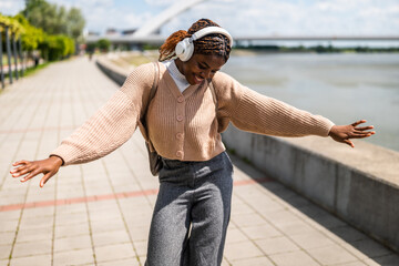 Happy young black woman is walking on foot path, listening music and smiling. She is enjoying sunny day outdoor.