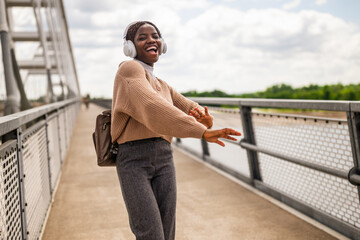 Happy young black woman is walking on bridge foot path, listening music and smiling. She is enjoying sunny day outdoor.