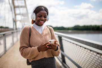 Happy young black woman is typing on her phone and smiling. She is enjoying sunny day outdoor and listening music while using smartphone.