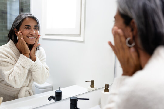 Mature happy woman touching and examining facial skin looking in bathroom mirror. - Powered by Adobe
