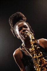Portrait of young adult Black woman playing saxophone on stage, wearing jewelry, looking into...