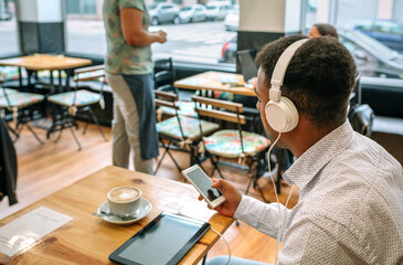 Unrecognizable young businessman wearing headphones and using smartphone while drinking cappuccino...