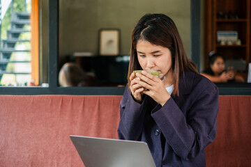 A focused young woman sips a hot beverage while working on her laptop in a stylish, cozy cafe...