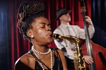 Black young adult woman playing saxophone with eyes closed in foreground, Caucasian young adult man...