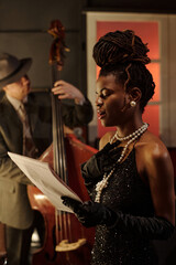 Young Black woman singing while holding sheet music, wearing elegant gloves and jewelry, standing...