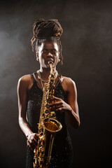 Black young woman playing saxophone on stage with eyes closed, wearing jewelry, standing against...