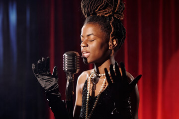 African American young adult woman singing into vintage microphone with eyes closed, wearing gloves...