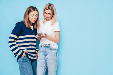 Two young beautiful smiling brunette hipster female in trendy summer clothes. Carefree women posing near blue wall. Positive models having fun. Cheerful and happy. Holding smartphone. Using apps
