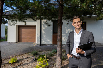 Real estate agent with clipboard and hand in pocket standing in front of modern house